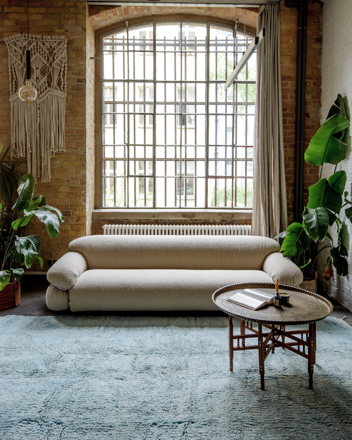 Beni Mrirt rug in finely striped light blue and cream, laying on floor in front of a cream curved couch and a large window. There are indoor plants to each side and a vintage morrocan table made of brass and wood.